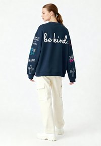 Jūras spēku liela izmēra svārki ar uzrakstu "būt laipnam." skriptā. Satur krāsainas grafikas un tekstu uz piedurknēm. Apvienots ar krēmkrāsas kravas biksēm.