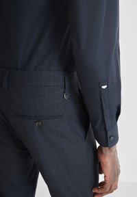 Muškarac nosi tamnoplave karirane hlače s stražnjim džepom na dugme i mornarskoplavu košulju s dugim rukavima i rukavima na dugme, ruka mu je opušteno pokraj tijela.