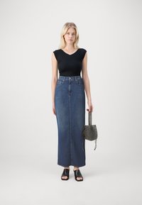 Jupe maxi en denim bleu foncé, ajustée à la taille, avec une coupe droite. Associée à un haut noir à manches courtes et des mules noires ouvertes.