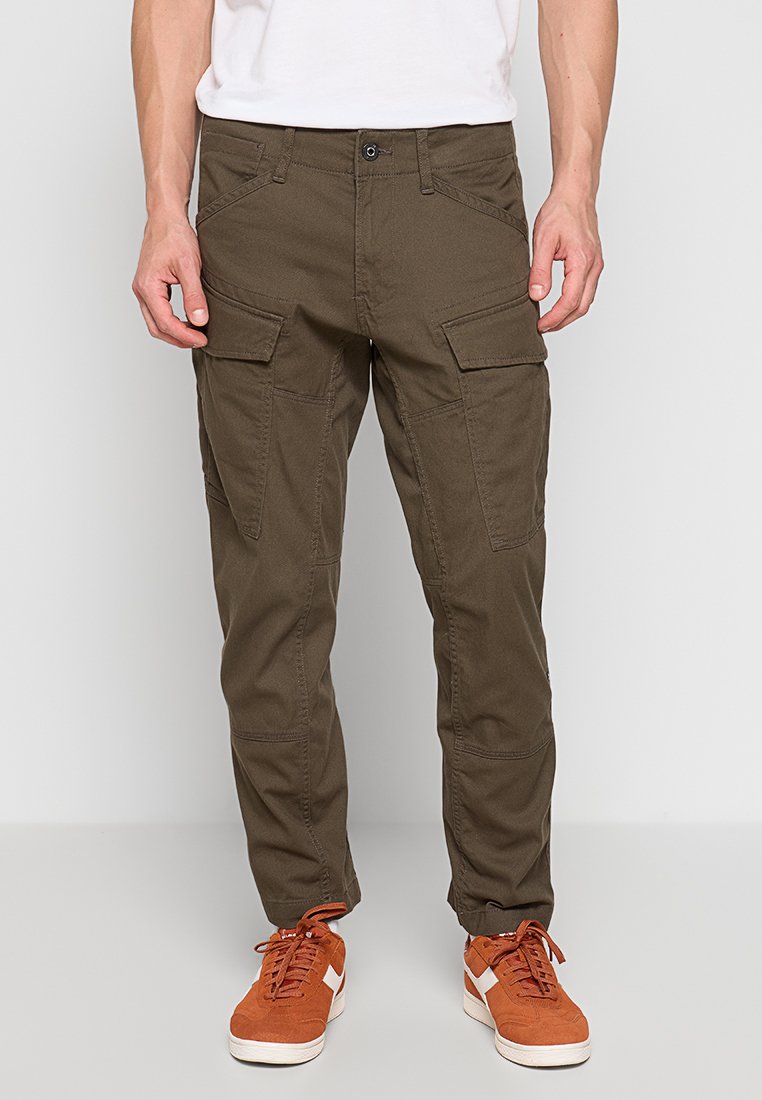Pantalones cargo de color verde oliva, hechos de algodón, con múltiples bolsillos frontales, un corte relajado y una pierna ligeramente ajustada. Llevados con zapatillas deportivas naranjas.