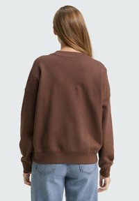 Brązowa bluzka z luźnym krojem, z prążkowanym okrągłym dekoltem i ściągaczami na rękawach. Tkanina wydaje się miękka z minimalnymi detalami. Połączona z niebieskimi dżinsami.