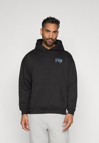 Férfi fekete kapucnis pulóverben, kék "City" logóval a mellkasán és világosszürke melegítőnadrágban, egy sima háttér előtt áll.