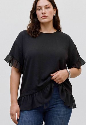 Femme portant un haut noir à manches courtes avec bords volantés et un jean bleu, accessoirisée avec un bracelet et des boucles d’oreilles en argent.