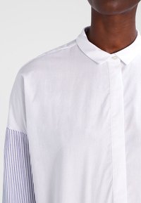 Bílá košile s knoflíky a kontrastním modro-bílým pruhovaným rukávem. Disponuje klasickým límcem a hladkým, lehkým materiálem.