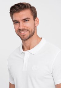 Biała koszulka polo wykonana z teksturowanej tkaniny, z zapięciem na dwa guziki oraz subtelnie wyszywanym logo po lewej stronie klatki piersiowej.