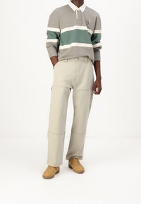 Mężczyzna w beżowych spodniach typu straight-leg, beżowych sneakersach i długim rękawie koszulce polo w szare, zielone i białe poziome paski.