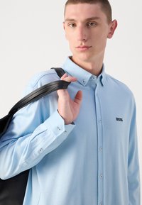 Lys blå button-up skjorte med en teksturert finish og svart "BOSS"-logo. Modell holder en svart veske over skulderen.