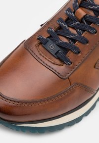 Zapatilla de cuero marrón con cordones azul marino y un acabado texturizado. Presenta un logo en la lengua y una entresuela blanca en contraste con una suela exterior con dibujo.