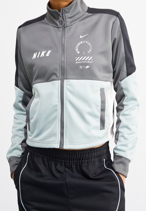 Persona que lleva una chaqueta Nike con cremallera gris y azul claro con logo y texto, combinada con pantalones negros con ribete blanco, manos en los bolsillos.