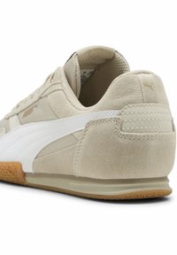Beżowe zamszowe sneakersy z białymi akcentami, teksturowana gumowa podeszwa i wyściełany kołnierz. Wyraźne logo Pumy w pobliżu pięty oraz unikalne detale przeszycia.