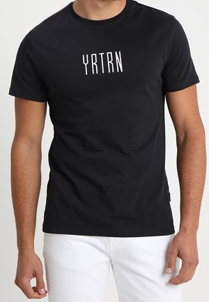 T-shirt noir à manches courtes avec texte blanc "YRTRN" sur la poitrine, porté par une personne à la peau claire et portant un pantalon blanc.