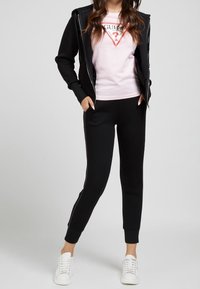 Svart zip-up hoodie över en ljusrosa T-shirt med en grafisk logotyp. Svarta joggerbyxor med sidostrepor, parat med vita sneakers.