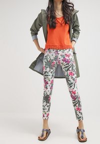 Olivově zelená voděodolná bunda, oranžový top s krajkovým lemem, květinové legíny v bílé, růžové a zelené barvě, doplněné tmavými sandály.