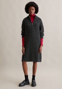 Donkergrijze ribgebreide jurk met een halve rits kraag, voorzien van lange mouwen en een relaxte pasvorm, gedragen over een rode turtleneck. Zwarte loafers dragen.