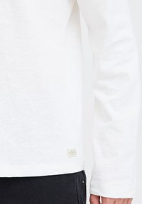 Weißes Langarmshirt mit weicher Textur, abgerundetem Saum und einem kleinen Etikett an der Unterseite. Schwarze Hose teilweise sichtbar.