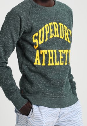 Sweat-shirt vert texturé avec graphique jaune "SUPERDRY ATHLETE". Présente des poignets et un ourlet côtelés ; associé à un short rayé clair.
