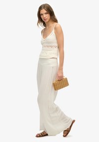 Top de crochet crema con tirantes finos, combinado con pantalones blancos ligeros y fluidos, sandalias marrones de tiras y un pequeño bolso de mimbre.