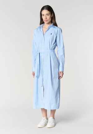 Donna che indossa un abito camicia lungo a righe verticali blu e bianche con cintura in vita e sneakers bianche, in piedi contro uno sfondo chiaro uniforme.