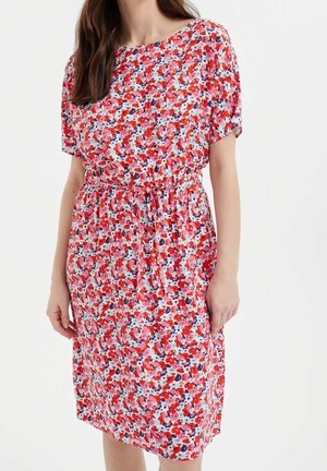 Frau trägt ein knielanges Kleid mit kurzen Ärmeln, das ein Blumenmuster in Rot, Pink, Weiß und Marineblau hat und mit einem taillierten Bindegürtel versehen ist.