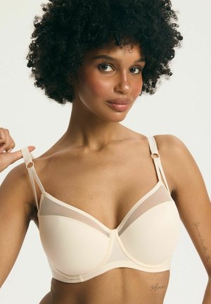 Kobieta o kręconych włosach regulująca ramiączko beżowego biustonosza z siateczkowym detalem na jednolitym jasnym tle.