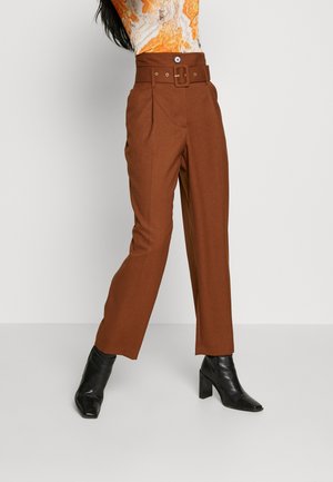 Woman wearing high-waist brown trousers with a belt, paired with black heeled ankle boots, standing against a plain background.