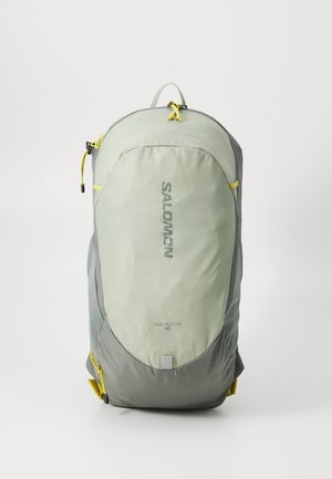 Sac à dos vert clair avec des accents gris, en tissu texturé, et un logo. Comprend des tirettes de zip jaunes et des poches latérales. Conçu pour une utilisation en extérieur.