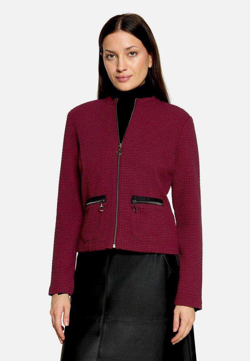 Femme aux cheveux lisses et foncés portant une veste zippée violette texturée avec des poches zippées noires et une jupe noire.