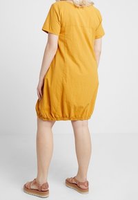 Senapsgul, kortärmad klänning med en samlad nederdel. Tillverkad av lättviktig tyg, har en subtil textur med en avslappnad passform, kombinerad med tan sandaler.