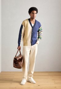 Sweater med blandede teksturer i beige, marineblå og mønstrede ærmer; cremefarvede bukser; læderbrun taske; hvide sneakers, trægulv i baggrunden.