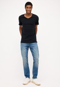 Tricou negru cu decolteu în V, mâneci scurte, asortat cu blugi albaștri deschiși și adidași albi. Modelul stă cu mâinile în spate pe un fundal simplu.