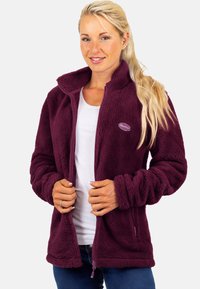 Vrouw met lang blond haar draagt een bordeauxrode fleecejas met ritssluiting over een wit shirt en blauwe spijkerbroek, terwijl ze met beide handen de jasranden vasthoudt.