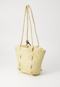 Wendbare Strohtasche in hellem Beige mit geflochtenen Griffen, dekorativen Knoten und einem geräumigen, schmalen Design.