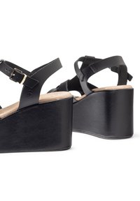 Des sandales compensées noires présentent des sangles en cuir croisées, une boucle et une semelle en liège. Le talon compensé est large et texturé, offrant une stabilité.