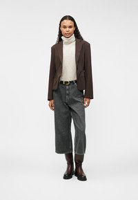 Brūns bleizers virs gaiša turtlenecka, apvienots ar brīviem pelēkiem džinsiem un līdz ceļgaliem ziliem brūnajiem zābakiem. Vienkārša fona dēļ izceļas apģērba detaļas.