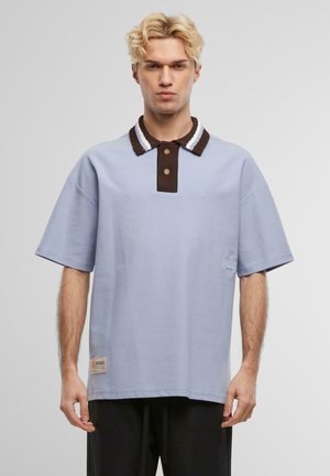 Polo-Shirt aus hellblauem Baumwollstoff mit einem braunen und weißen Streifenkragen, einer Zwei-Knopf-Leiste und einem seitlichen Etikett-Patch-Detail.