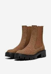 Bottines en suède marron avec panneaux élastiques sur les côtés et une semelle en caoutchouc noir chunky, présentant un bout rond et une finition texturée.