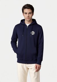 Felpa con zip color navy realizzata in tessuto morbido, dotata di logo frontale, cappuccio con cordino e tasche a marsupio. Polsini e orlo a coste.