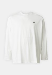 Vit långärmad tröja med rundad hals och en liten grön krokodil-logotyp på vänster bröst, visad mot en enkel bakgrund.