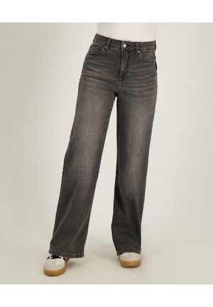 Persoon draagt een high-waisted, wijd uitlopende, vervaagde zwarte spijkerbroek en witte sneakers met lichtbruine zolen, staand op een effen achtergrond.