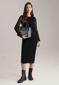 Schwarzes knielanges Kleid mit langen durchsichtigen Ärmeln, kombiniert mit einer marineblauen Handtasche mit Blumenmustern und einem Logo. Das Modell trägt schwarze Schnürstiefel.