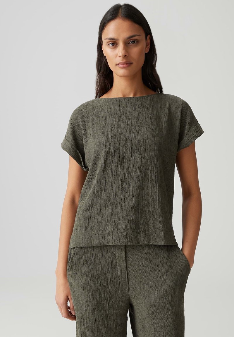 Woman wearing textured olive green short-sleeve top and matching high-waist pants, standing with one hand in pocket against plain background.