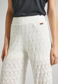 Pantalones de punto de ganchillo blancos que presentan un patrón de diamantes, tela texturizada y una cinturilla lisa con una pequeña etiqueta de cuero marrón.