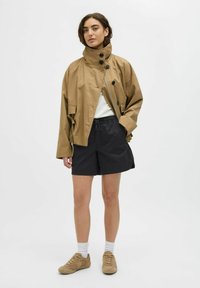 Chaqueta impermeable marrón y de gran tamaño con un cuello alto y botones negros, combinada con pantalones cortos negros y zapatillas beige. Calcetines blancos visibles.