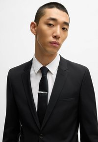 Costume noir avec un revers cranté, chemise habillée blanche et cravate noire avec une pince à cravate en argent. Tissu lisse et design ajusté.