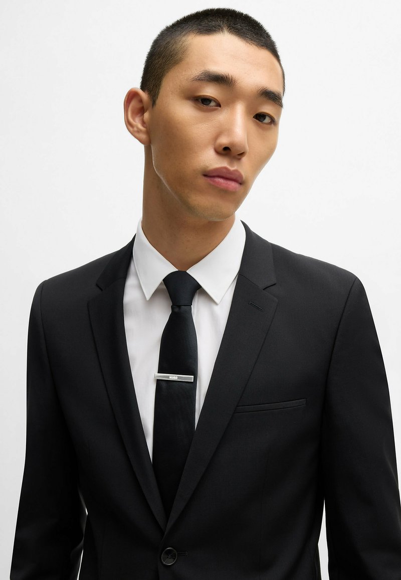 Costume noir avec un revers cranté, chemise habillée blanche et cravate noire avec une pince à cravate en argent. Tissu lisse et design ajusté.