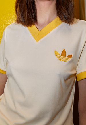 Frau trägt ein cremefarbenes Adidas-Shirt mit gelbem Besatz am Kragen und an den Ärmeln sowie einem gelben Adidas-Logo auf der linken Brust.