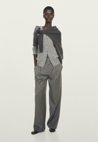 Persona alta con un cardigan grigio a bottoni, un maglione grigio scuro drapeato sulle spalle, pantaloni grigi a righe sottili comodi e scarpe nere.