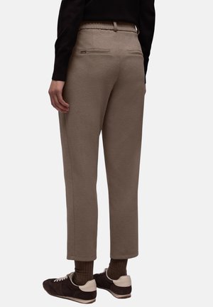 Beżowe spodnie dresowe wykonane z miękkiego materiału, z nogawkami zwężanymi ku dołowi, elastycznym pasem i dwoma tylnymi kieszeniami. Noszone z ciemnymi butami i skarpetkami.