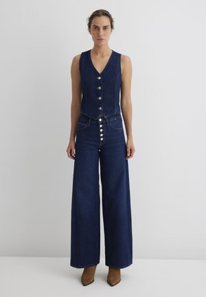 Denim jumpsuit with a sleeveless, fitted top featuring a deep V-neck and five large buttons. Wide-leg trousers in dark blue, textured fabric.