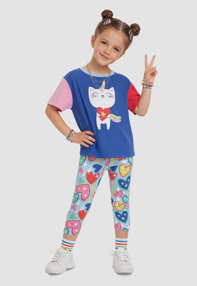 Niña joven con peinado de moños dobles, vistiendo una camiseta colorida con tema de gatos y leggings con estampado de corazones, mostrando el signo de la paz con su mano derecha.